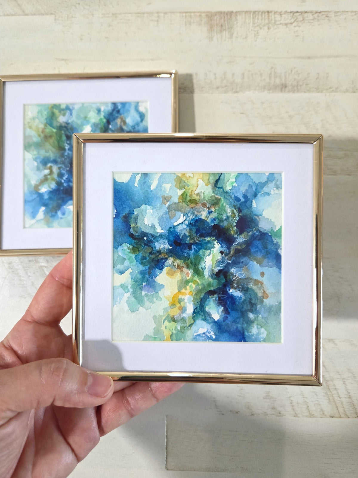 A hand holds a small square framed abstract watercolor print with blue, green, and yellow tones, white matting, and a thin gold frame. Another matching framed print is blurred in the background against a light wood wall.